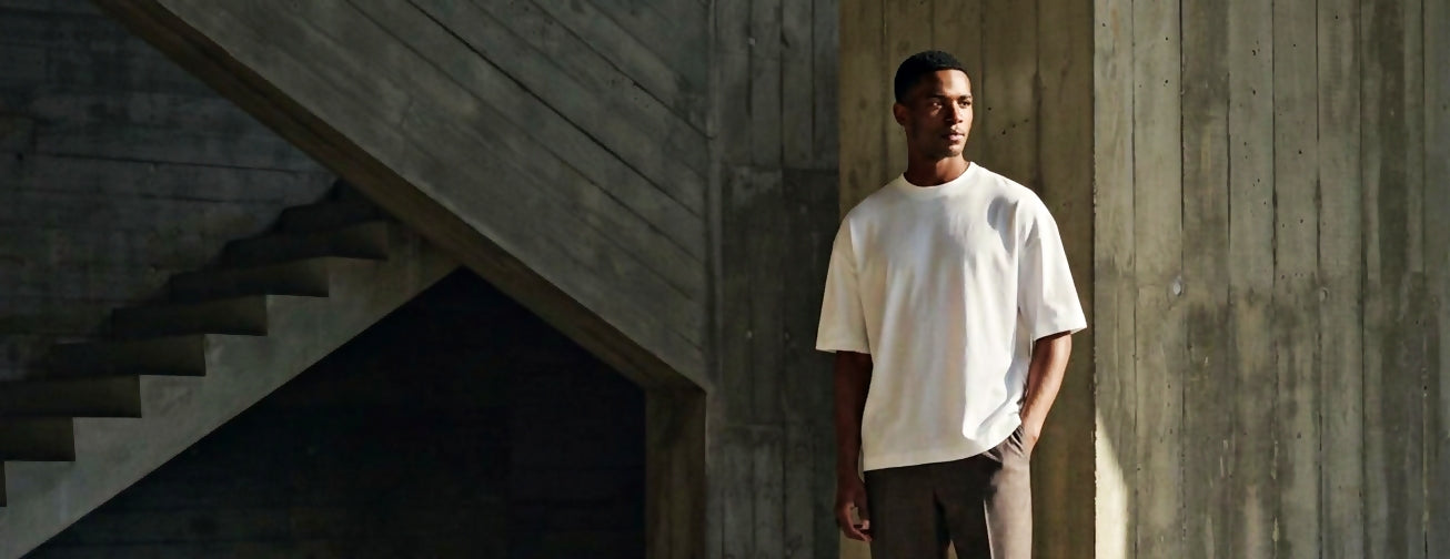 A man wearing an oversized white WILClothing blank crewneck t-shirt and dark gray pants, standing in a modern concrete structure with a staircase on the left.